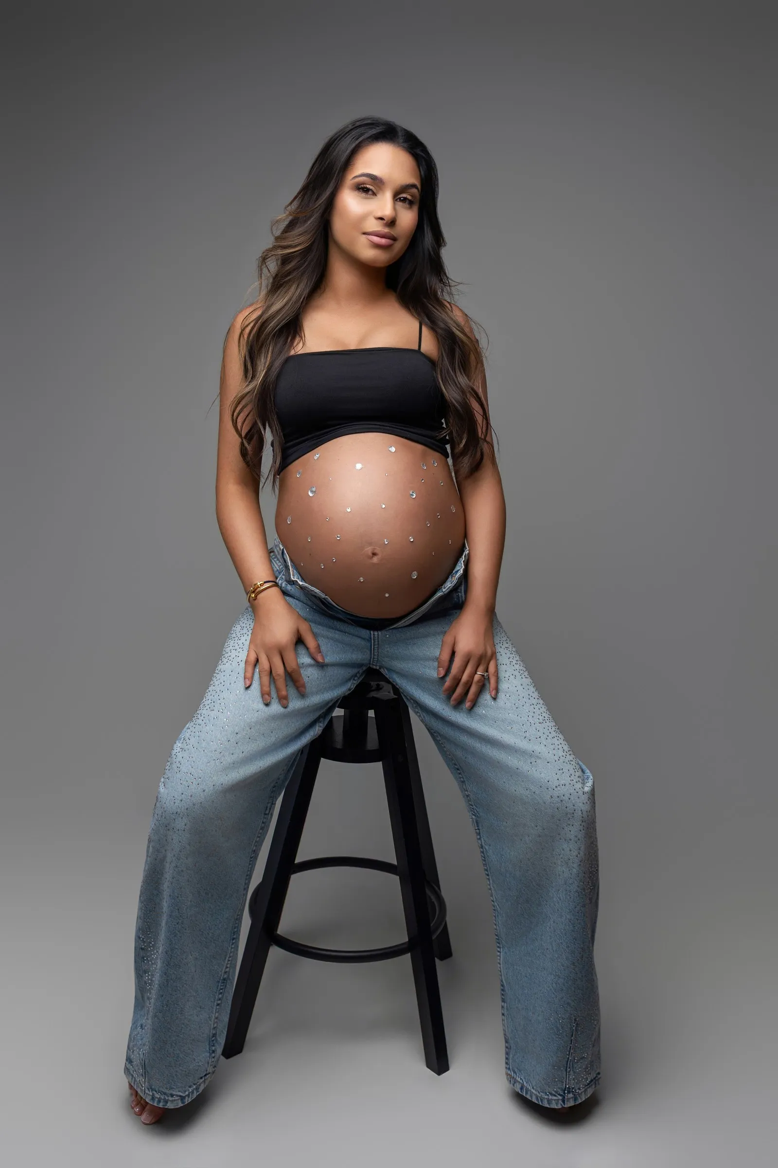 Future maman en jean strass, bijoux sur le ventre, assise sur un tabouret en studio près de Toulouse