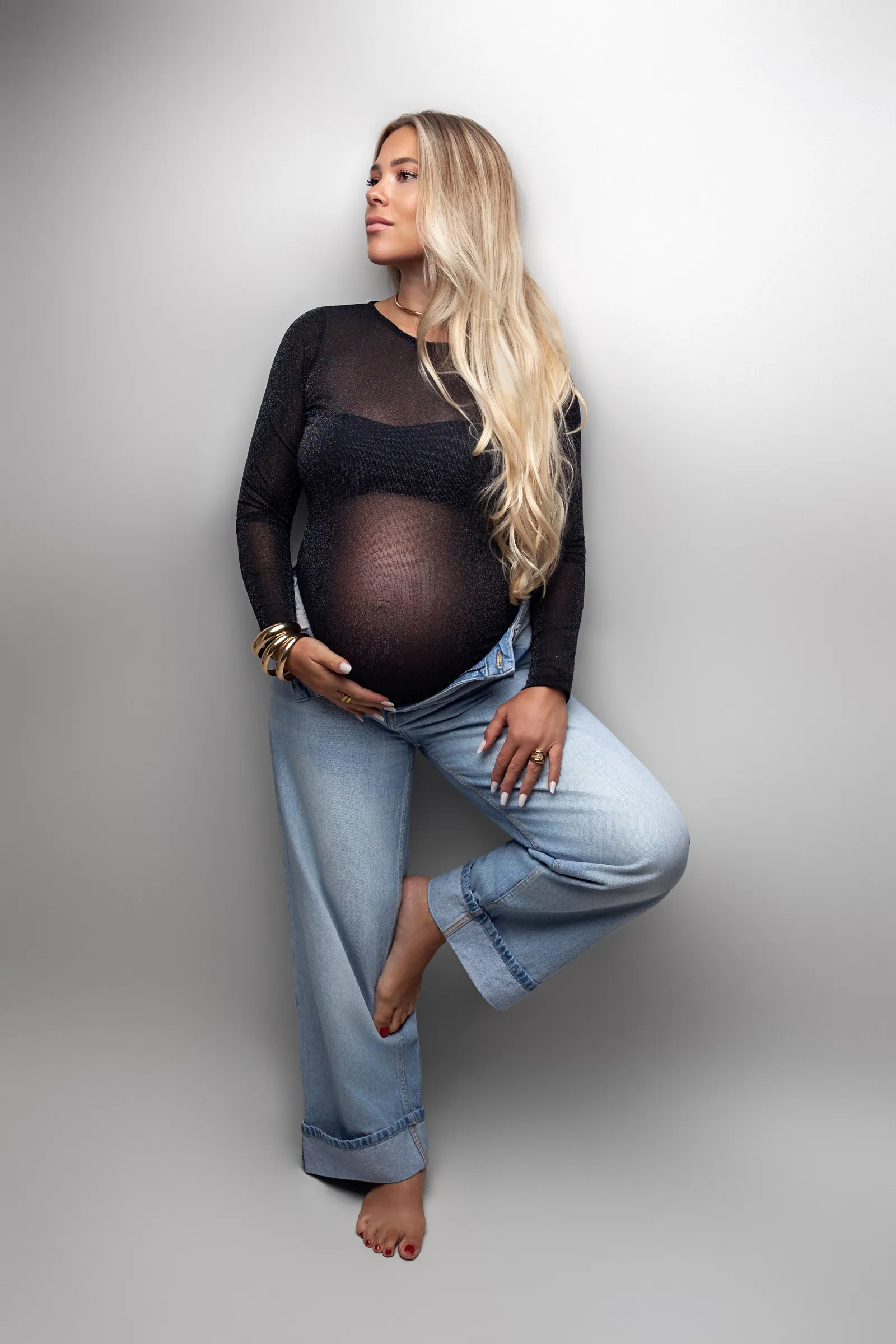 Future maman en top transparent noir et jean large, adossée à un mur clair, séance photo grossesse en studio près de Toulouse